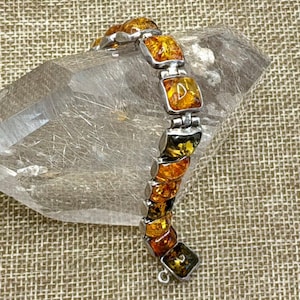 May include: A link bracelet featuring square-shaped, translucent amber stones in varying shades of honey, orange, and dark brown, set within silver-toned metal frames. The bracelet includes a thin silver chain extension with a small circular clasp.