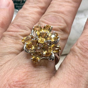 May include: A silver ring featuring a cluster of oval-cut, golden-yellow gemstones. The gemstones are arranged in a floral pattern, creating a sparkling and eye-catching design. The ring is set on a hand, showcasing the jewelry's size and style.