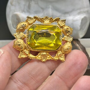 May include: A vintage gold-toned brooch featuring a large, rectangular, yellow gemstone at its center. The gemstone is surrounded by an ornate, gold-colored frame with intricate detailing. The brooch is held in a hand.