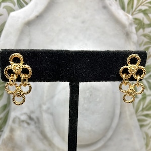Peut inclure: Une paire de boucles d'oreilles dorées avec un motif floral. Chaque boucle d'oreille présente trois grappes en forme de fleur reliées, avec un motif texturé en forme de corde. Les boucles d'oreilles sont exposées sur un présentoir noir.