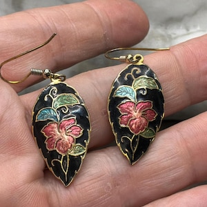 May include: A pair of leaf-shaped earrings with a black background, featuring a red and pink flower design with green and blue accents. The earrings have gold-colored hooks.