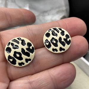 May include: A pair of round earrings with a cream-colored base and a black leopard print design. The earrings are held in a person's hand, showcasing their size and detail. The earrings are a fashion accessory.