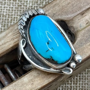 Peut inclure: Une bague en argent ornée d'une grande pierre turquoise bleu vif, lisse et de forme ovale, sertie dans une monture en métal poli. L'anneau est décoré de motifs de feuilles gravés d'un côté et de petites sphères en argent de l'autre.