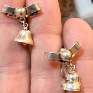 以下が含まれることがあります： 銀色のイヤリングのペア。各イヤリングは、中央に小さなボールが付いた弓形のトップと、ぶら下がっているベルチャームが特徴です。イヤリングは人の手で持たれています。