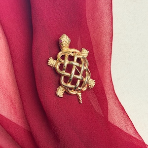 May include: A gold-colored turtle brooch with a woven shell design and a small, red gemstone eye. The brooch is set against a backdrop of a deep red, sheer fabric.