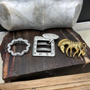 May include: Three decorative brooches on a dark wooden surface. A silver-toned, scalloped-edge brooch, a silver-toned square brooch with paw prints and a speech bubble that says "MEOW!", and a gold-toned zebra brooch.