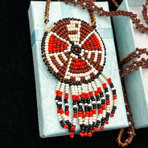 May include: A beaded necklace with a circular pendant featuring a geometric design in red, white, brown, and black beads. The pendant has beaded fringe in red, white, and black. The necklace is displayed on a light blue box.