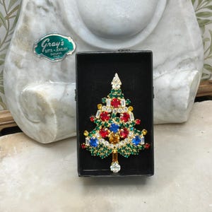 May include: A Christmas tree brooch with a gold-tone metal base, adorned with red, blue, yellow, and clear rhinestones. The brooch is displayed in a black box. The brooch is a festive holiday accessory.