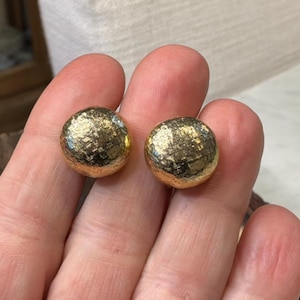 May include: A pair of round, gold-colored earrings with a textured surface. The earrings have a metallic sheen and a domed shape. The jewelry is likely made of metal and is suitable for everyday wear or special occasions.