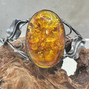 May include: A silver cuff bracelet featuring a large, oval-shaped, translucent amber stone in the center. The metal band is decorated with detailed, sculpted silver leaves on both sides of the central amber setting, creating a botanical motif.