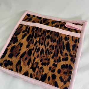 May include: A square, quilted pot holder with a leopard print pattern in shades of brown and black. The edges and a horizontal stripe are trimmed with a light pink fabric. A small pink loop is attached to the top edge.