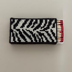May include: A rectangular matchbox cover featuring a black and white cross-stitch zebra stripe pattern. The cover is crafted from yarn and fits over a standard cardboard matchbox containing wooden matches with red tips visible on the right side.