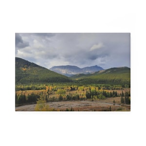 May include: A landscape canvas print featuring a mountain range under a cloudy sky. The foreground shows a valley with trees in shades of green and yellow. The mountains are in the background.