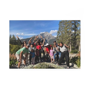 Puede incluir: Un grupo de personas posa para una foto al aire libre. Están de pie frente a una montaña y árboles. El cielo es azul con algunas nubes. Algunas personas llevan sombreros y gafas de sol.