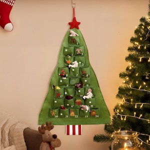 Pode incluir: Um calendário do advento em forma de árvore de Natal de feltro verde com 24 bolsos numerados, cada um contendo uma pequena figura festiva. A árvore é decorada com uma estrela vermelha e está pendurada numa parede.