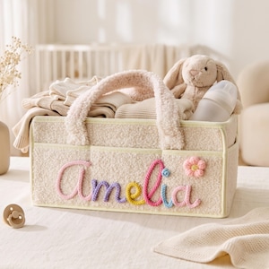 May include: A cream-colored sherpa storage basket with two handles, featuring the name Amelia embroidered in multi-colored yarn and a small pink flower appliqué. The basket is filled with folded beige clothing, a plush rabbit, and a baby bottle.