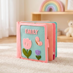 May include: A felt quiet book with a light blue cover featuring the name EMILY in pink letters, a large pink flower, a small purple flower, and a pink butterfly. The book has multiple pages in shades of pink and blue, constructed from soft felt fabric.