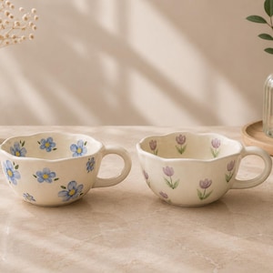 May include: Two ceramic mugs with a scalloped rim and a glossy cream finish. The left mug features hand-painted blue flowers with yellow centers, while the right mug displays small purple tulip illustrations with green stems and leaves.