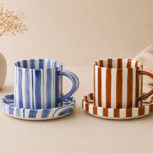 May include: Two ceramic mugs with matching saucers, one featuring vertical blue stripes and the other featuring vertical brown stripes. Each mug has a rounded handle and a cylindrical shape, with the stripes continuing onto the saucers.