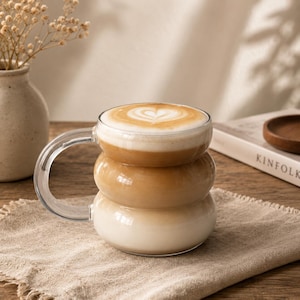 May include: A clear glass mug with a distinctive three-tiered, rounded bubble shape, featuring a large curved handle. The mug is filled with a layered coffee beverage topped with a white foam heart design, showcasing a gradient from dark brown to cream.