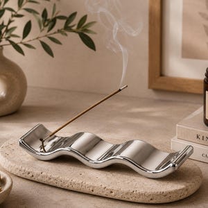 May include: A polished silver-toned wavy incense holder resting on a textured beige stone tray. A single incense stick is inserted into the holder, emitting a thin trail of white smoke. The tray is oval-shaped with a porous, natural surface finish.