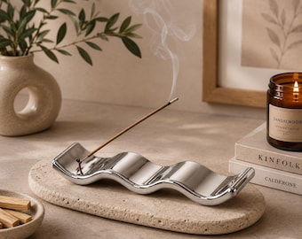 May include: A polished silver-toned wavy incense holder resting on a textured beige stone tray. A single incense stick is inserted into the holder, emitting a thin trail of white smoke. The tray is oval-shaped with a porous, natural surface finish.