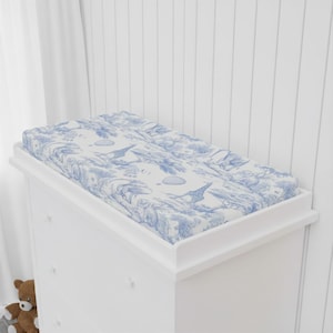 Op de afbeelding: Een witte commode met een blauw-wit patroon verschoonkussen. Het kussen heeft een toile-ontwerp met giraffen, bomen en andere elementen. De commode heeft een eenvoudig, strak ontwerp.