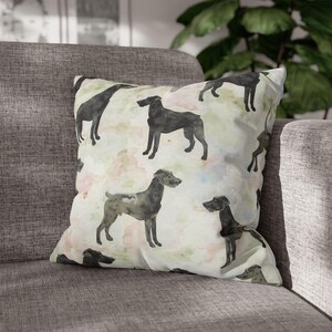 May include: Square cushion with a watercolour design of black dog silhouettes on a pale background with pastel colour splashes. The cushion is on a grey sofa.