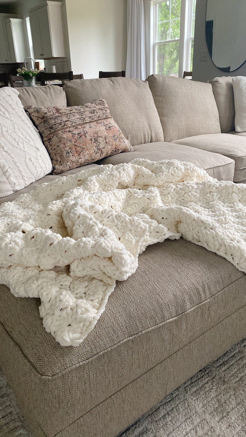 Pu&ograve; includere: Un divano angolare beige con una coperta di maglia bianca e un cuscino con un motivo. Il divano si trova in un soggiorno con una finestra e uno specchio sullo sfondo.