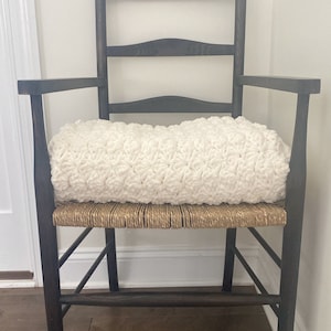 Pu&ograve; includere: Una sedia in legno scuro con una seduta in vimini intrecciato e un cuscino in crochet bianco. La sedia si trova in una stanza con pavimento in legno duro e un tappeto a motivi.
