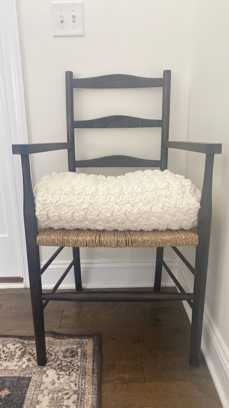 Pu&ograve; includere: Una sedia in legno scuro con una seduta in vimini intrecciato e un cuscino in crochet bianco. La sedia si trova in una stanza con pavimento in legno duro e un tappeto a motivi.