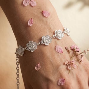 Peut inclure: Un bracelet argenté orné de motifs floraux ajourés et délicats. Le bracelet est composé de petites fleurs détaillées et est présenté sur un poignet, entouré de pétales de fleurs roses. Il possède une chaîne réglable.