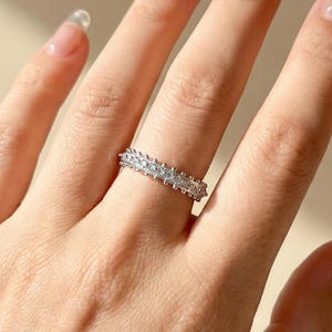 May include: A silver ring with a continuous row of small, clear gemstones. The ring is set on a hand with a neutral skin tone, and the ring's design is visible against the skin. The ring appears to be made of silver or a similar light-colored metal.