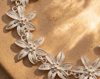 May include: A silver-colored floral necklace with delicate filigree detailing. The necklace features multiple flower-shaped pendants connected by small links. The flowers have five petals and a central bead. The necklace is displayed on a light brown surface.