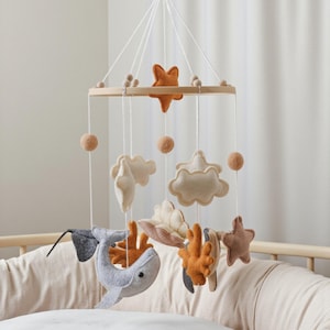 May include: A nursery mobile with felt sea-themed decorations, such as a whale, starfish, clouds, and coral. The mobile hangs from a wooden ring with strings and small felt balls. The colour scheme includes grey, orange, beige, and brown.