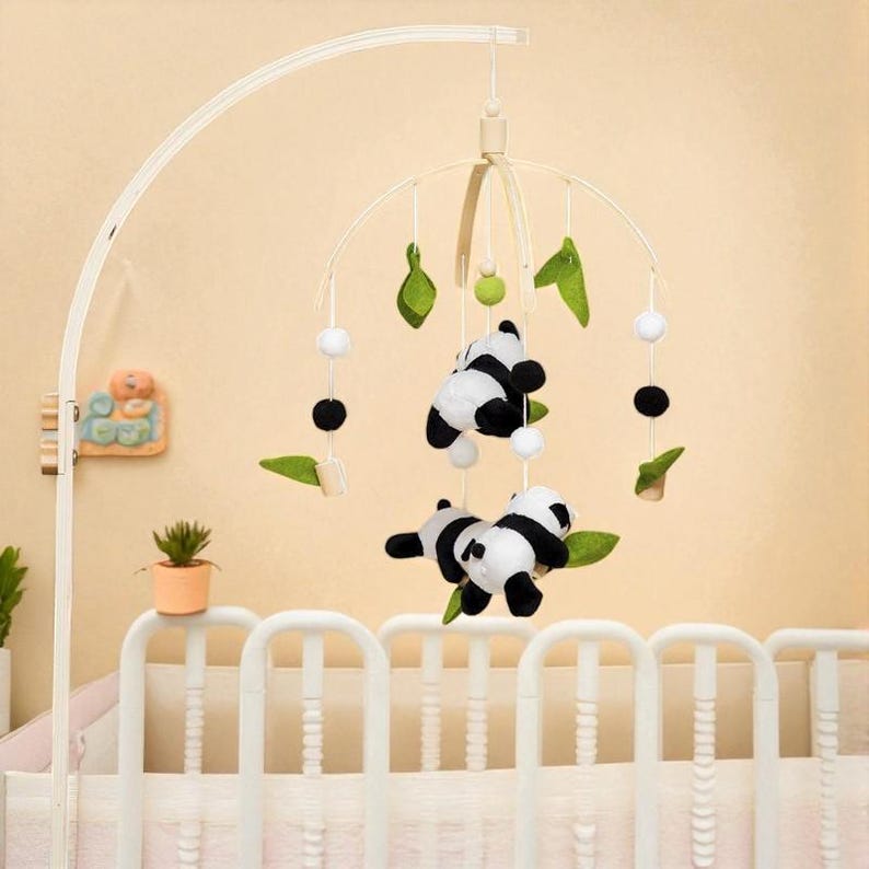 May include: A baby crib mobile with a cream-colored frame. The mobile features two plush panda bears, green leaf accents, and white and black spherical ornaments. The mobile is suspended above a white crib.