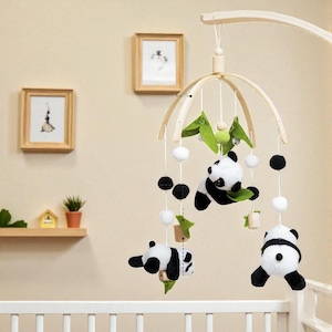 May include: A baby mobile featuring plush panda bears, green felt leaves, and white and black pom-poms. The mobile is suspended from a wooden frame and hangs above a white crib.