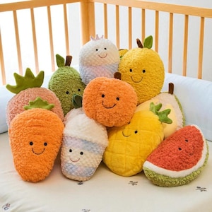 Puede incluir: Una colección de cojines de felpa con forma de fruta, que incluye una zanahoria, una fresa, una sandía, una piña y un limón. Cada cojín tiene una cara sonriente y está hecho de tela suave y texturizada. Los cojines están colocados sobre una superficie blanca.