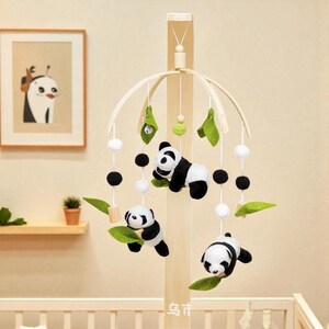 May include: A wooden baby mobile featuring three plush panda bears in black and white, suspended from a natural wood frame. The mobile includes green felt leaves, white and black pom-poms, and is designed for a nursery.