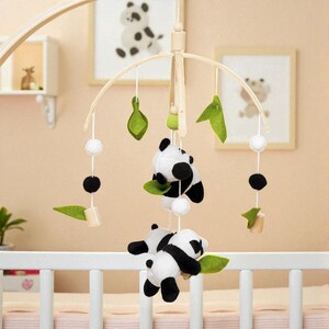 May include: A baby crib mobile featuring panda bear plush toys, green felt leaves, white pom-poms, and wooden accents. The mobile hangs from a wooden frame and is designed to be suspended above a crib. The background includes framed panda bear art.