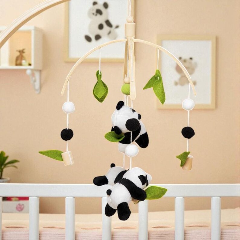 May include: A baby crib mobile featuring panda bear plush toys, green felt leaves, white pom-poms, and wooden accents. The mobile hangs from a wooden frame and is designed to be suspended above a crib. The background includes framed panda bear art.