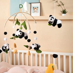 May include: A baby crib mobile featuring panda bears, green leaves, and black and white pom-poms. The mobile has a wooden frame and is suspended above a white crib. The pandas are black and white plush toys.