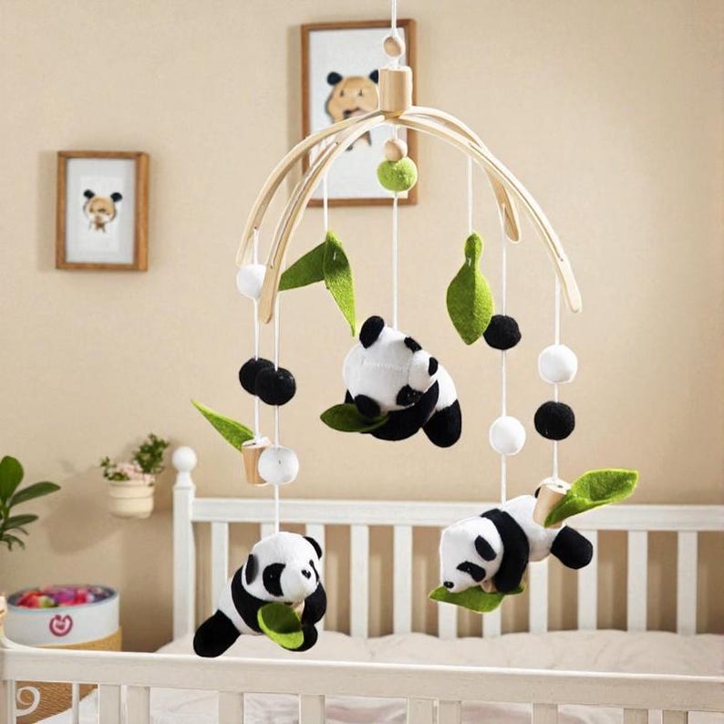May include: A baby mobile featuring panda bears, green leaves, and white and black pom-poms suspended from a wooden arch. The mobile hangs above a white crib. Two framed panda bear illustrations are visible in the background.