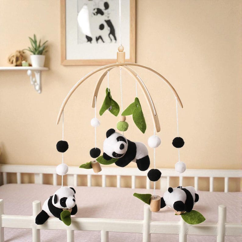 May include: A baby mobile featuring panda bears, green felt leaves, and white and black pom-poms. The mobile is suspended from a wooden arch and hangs above a white crib. A framed picture of a dog is visible in the background.
