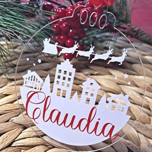 Può includere: Ornamento natalizio trasparente con una silhouette bianca di paesaggio urbano e il nome "Claudia" in corsivo rosso. Babbo Natale e le renne sono raffigurati sopra la città. L'ornamento è appeso con un nastro rosso.