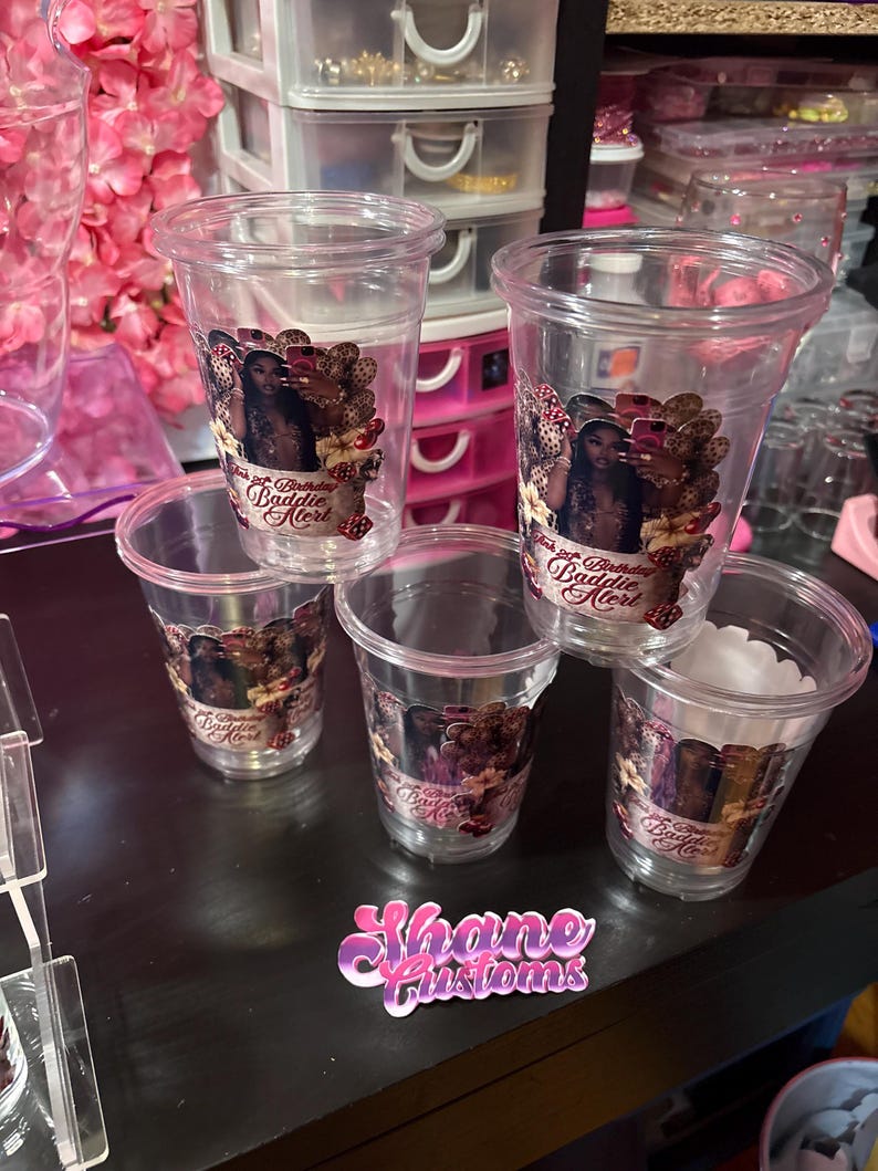 May include: A collection of clear plastic cups with a printed design. The design features a woman, text that reads "21st Birthday Baddie Alert", and decorative elements. A sticker with the text "Shane Customs" is also visible.