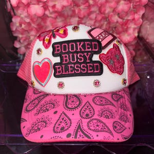 以下が含まれることがあります： 「BOOKED BUSY BLESSED」の文字が入った白いフロントパネルが付いたピンクのトラッカーハット。ピンクのペイズリー柄のつばと、ピンクのラインストーンと装飾パッチが特徴です。