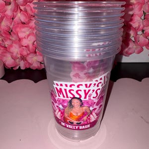 May include: A stack of clear plastic cups with a printed design. The design features the text "Missy's" and "M-Vally Bash" with a photo of a woman. The cups are stacked on a white coaster, with pink flowers in the background.