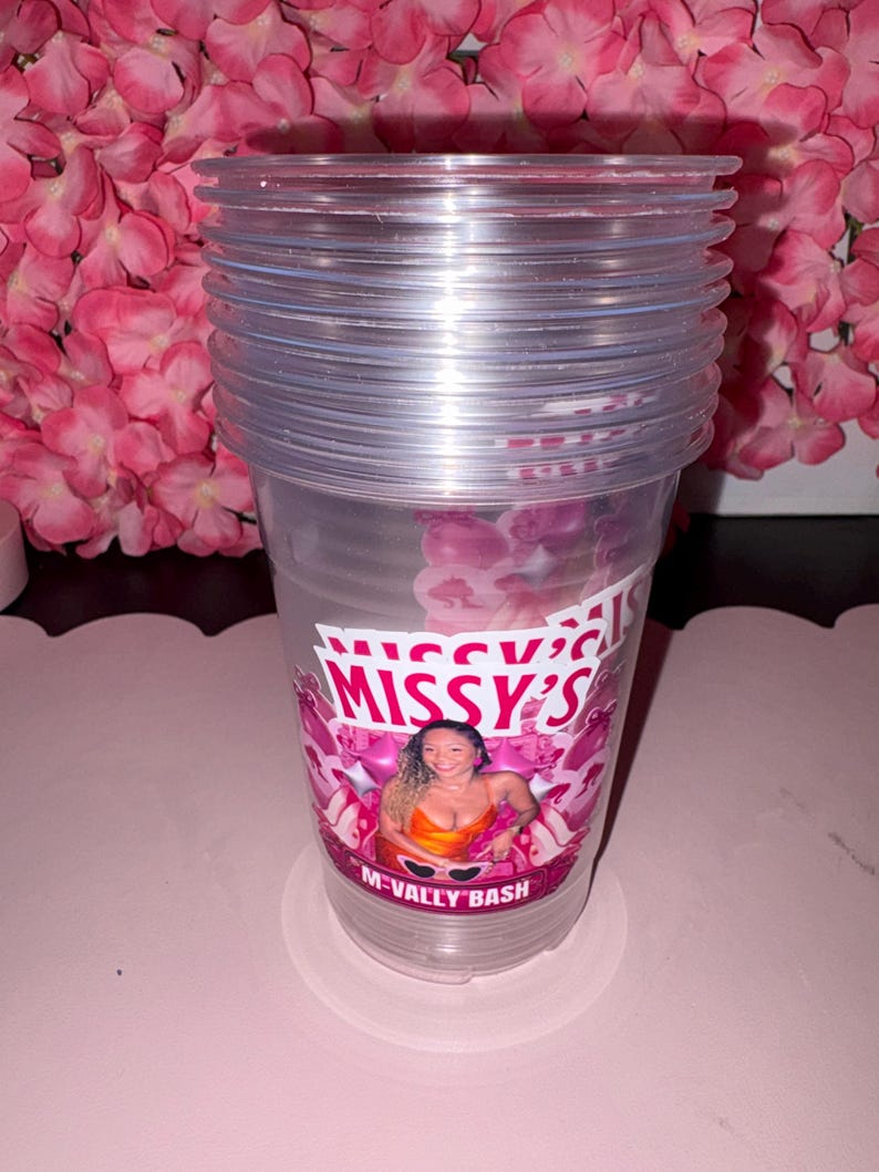 May include: A stack of clear plastic cups with a printed design. The design features the text "Missy's" and "M-Vally Bash" with a photo of a woman. The cups are stacked on a white coaster, with pink flowers in the background.