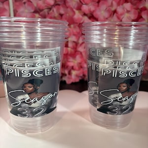 May include: Two clear plastic cups with a printed design. The design features the word "PISCES" and "Season 32" with an illustration of a woman. The background is a blurred display of pink flowers.